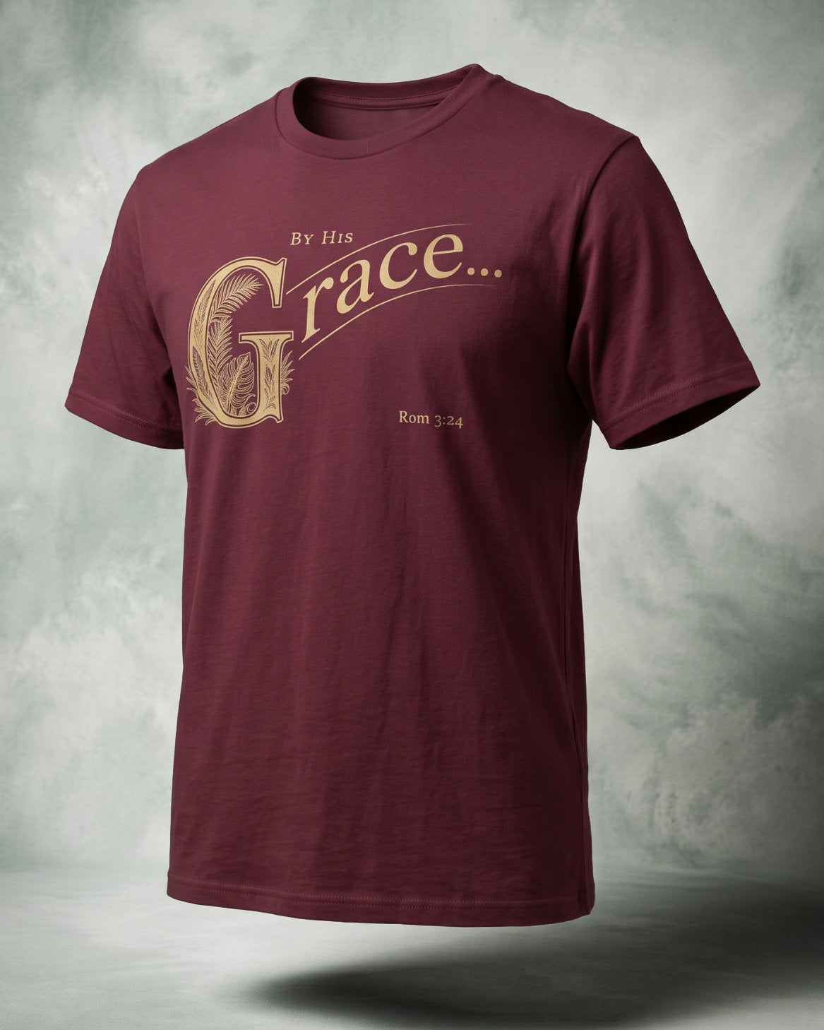 By His Grace t-shirt in maroon with gold ornamental lettering and Rom 3:24, three-quarter studio view
